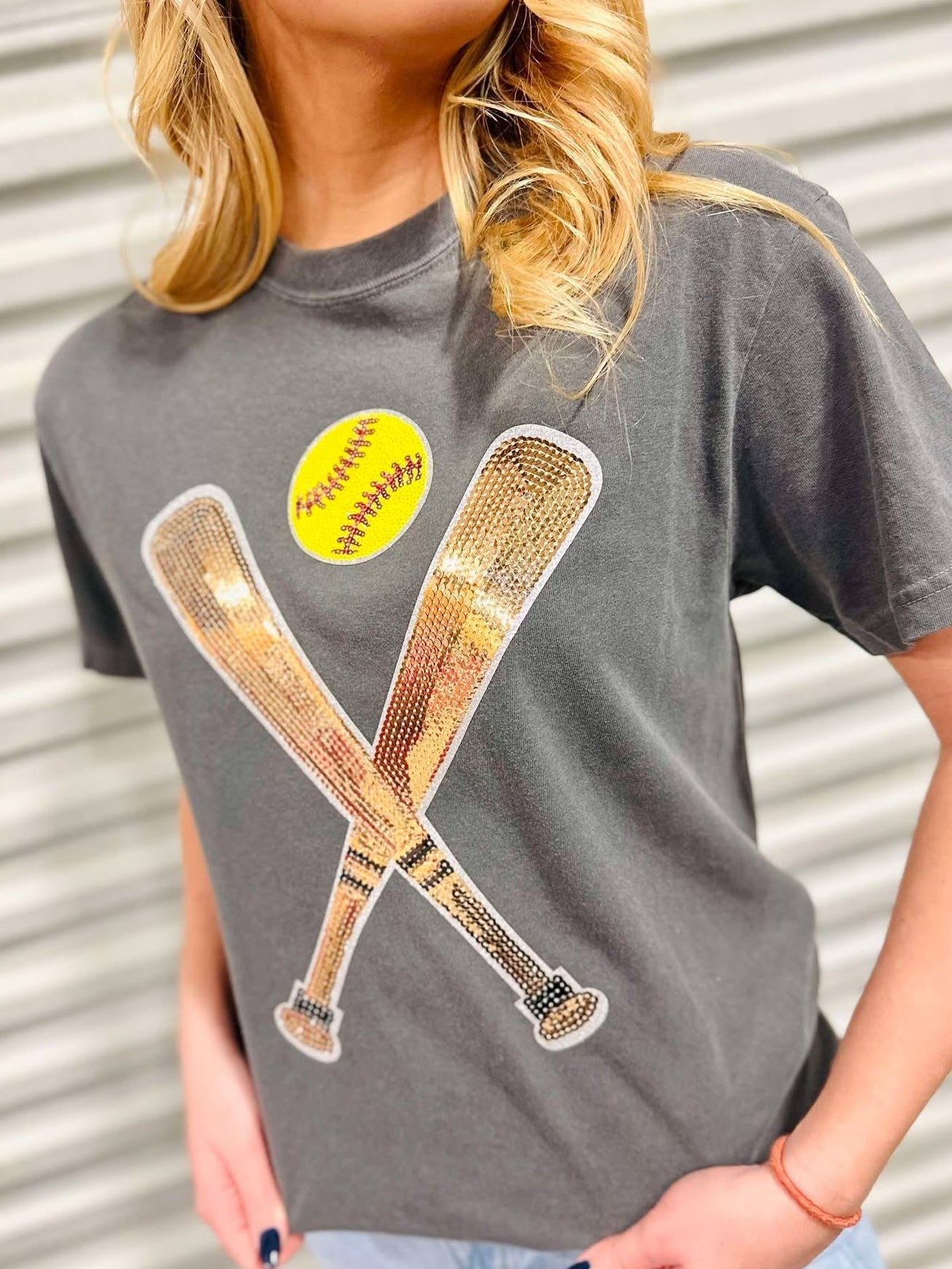 A female model wearing a gray t-shirt with a sequin patch on the front showing two softball bats and a yellow softball.