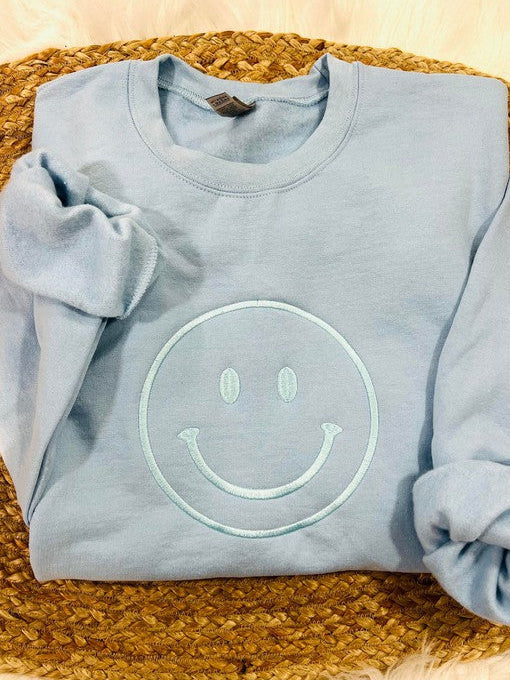 A light blue sweatshirt with an embroidered lettering of a smiling face on the chest, displayed on a woven mattress.