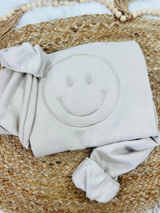 A beige embroidered sweatshirt with a happy face design on the front, displayed on a woven mat.