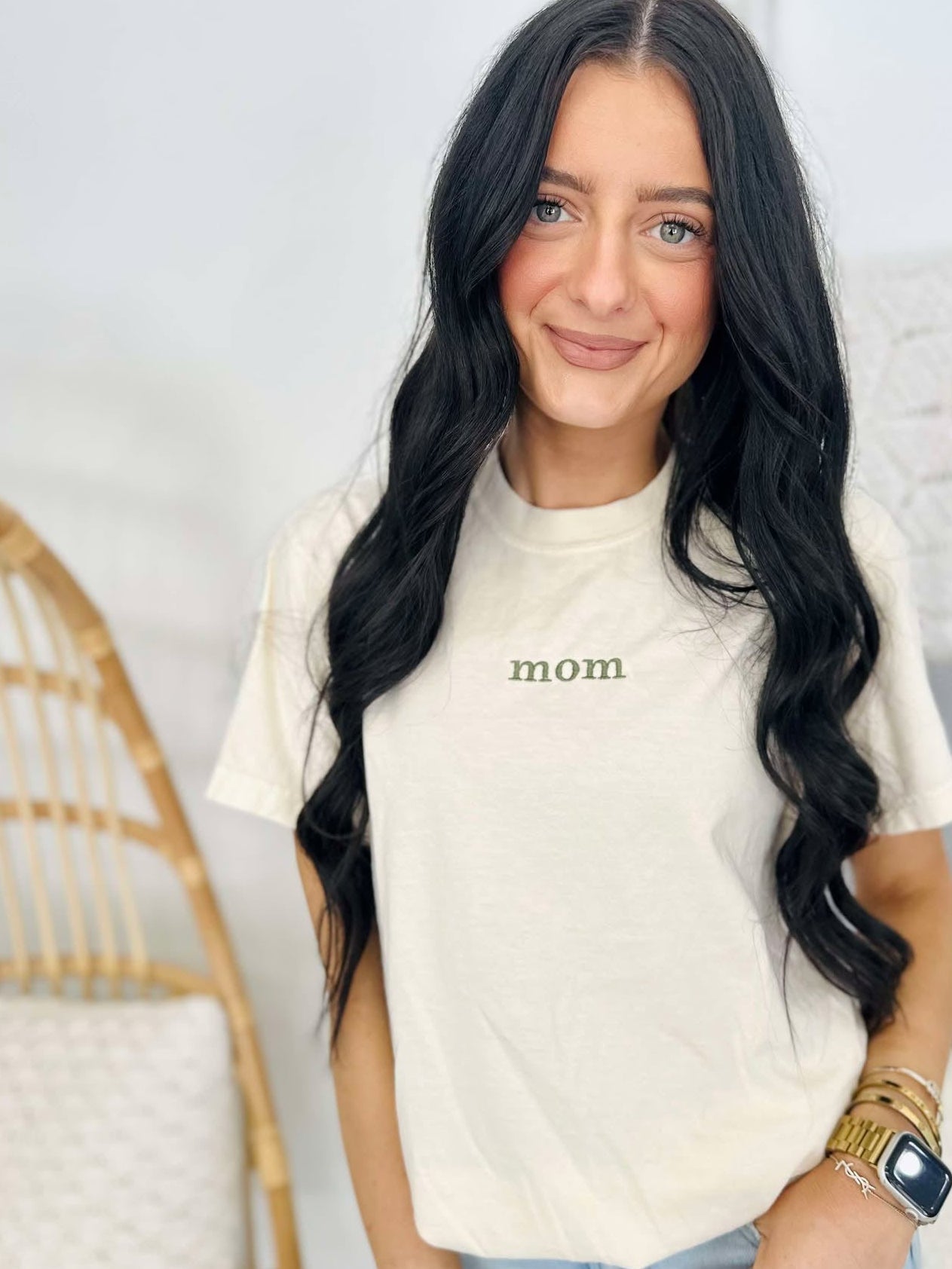 A woman wearing a white short-sleeved t-shirt with the word 'Mom' embroidered on the front, standing in a room with a wooden rocking chair and a watch on her wrist.