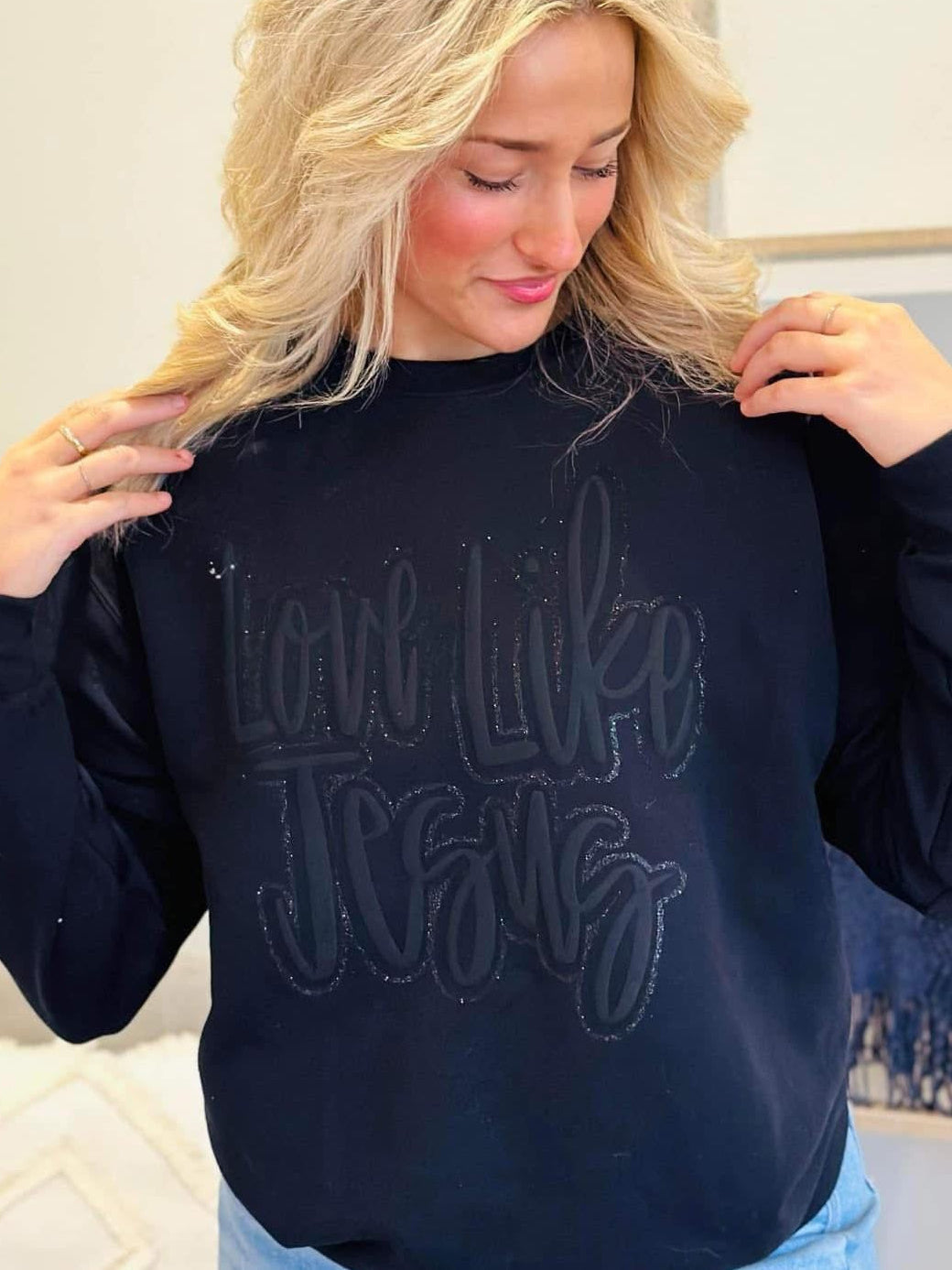 Woman wearing a black sweatshirt with Black monocromatic Love like Jesus , sitting on a couch.