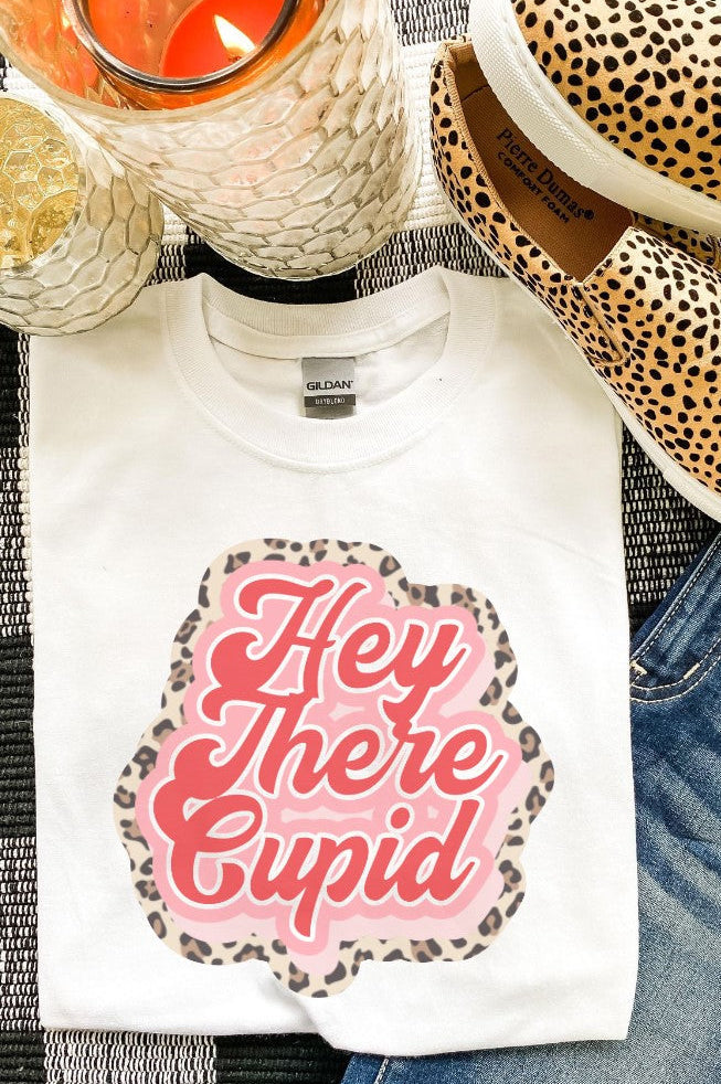 White t-shirt with 'Hey There Cupid' design, leopard print shoes, and candle on a checkered fabric background