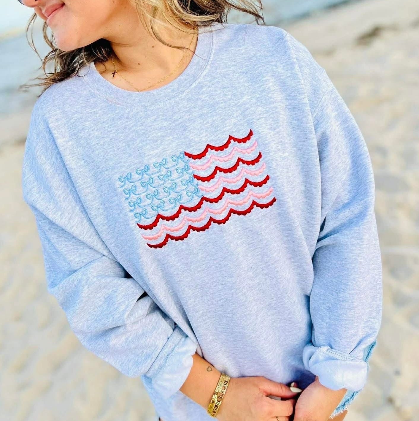 Person wearing a light blue sweatshirt with a colorful lace and bow American flag wave design on a beach.